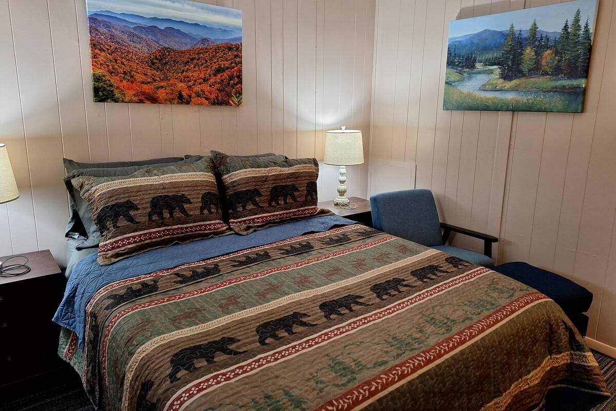 A hotel room with a bed, two pillows with bear designs, a blue chair, a nightstand, a lamp, and two paintings of landscapes.