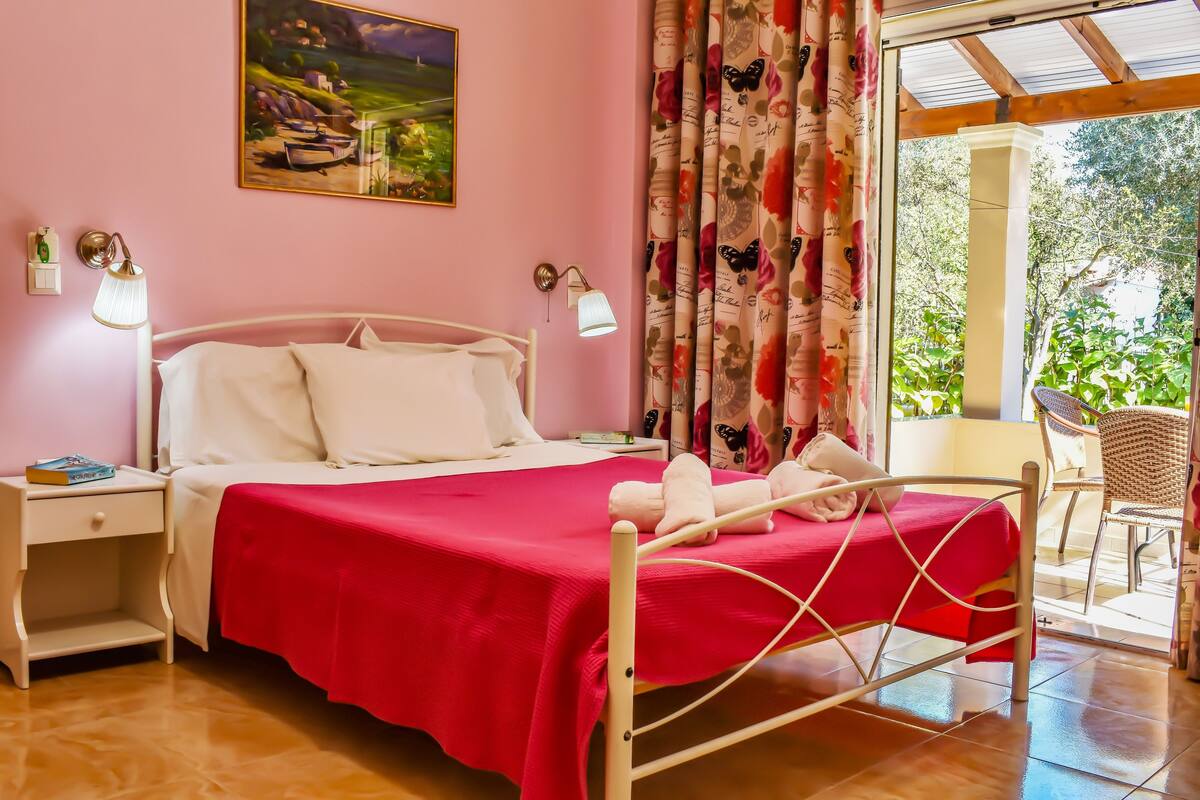 A bedroom with a bed, nightstands, a lamp, a painting, and a view of the outdoors.