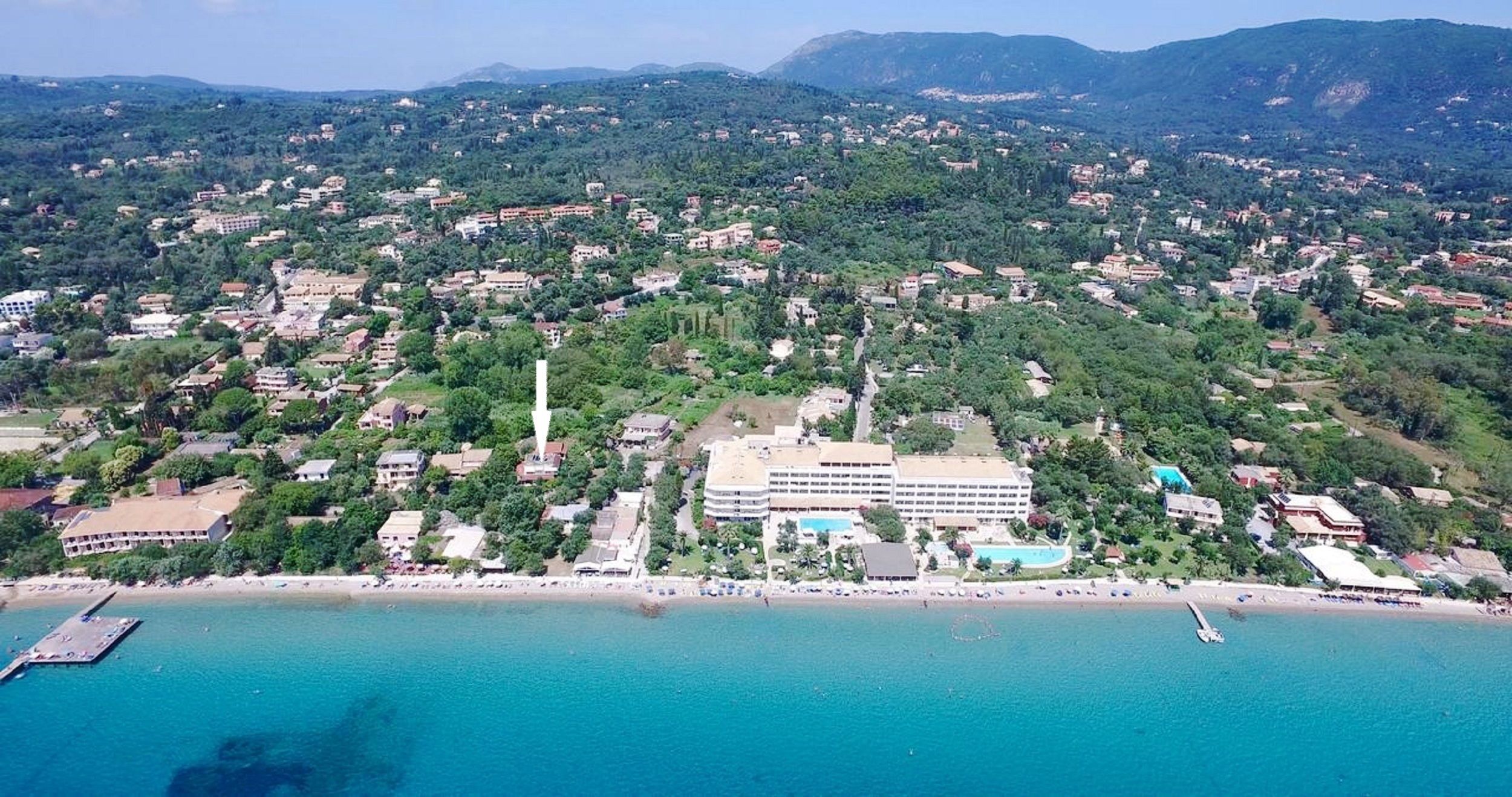 An aerial view of a coastal town with a prominent hotel featuring a swimming pool, surrounded by lush greenery and a beach.
