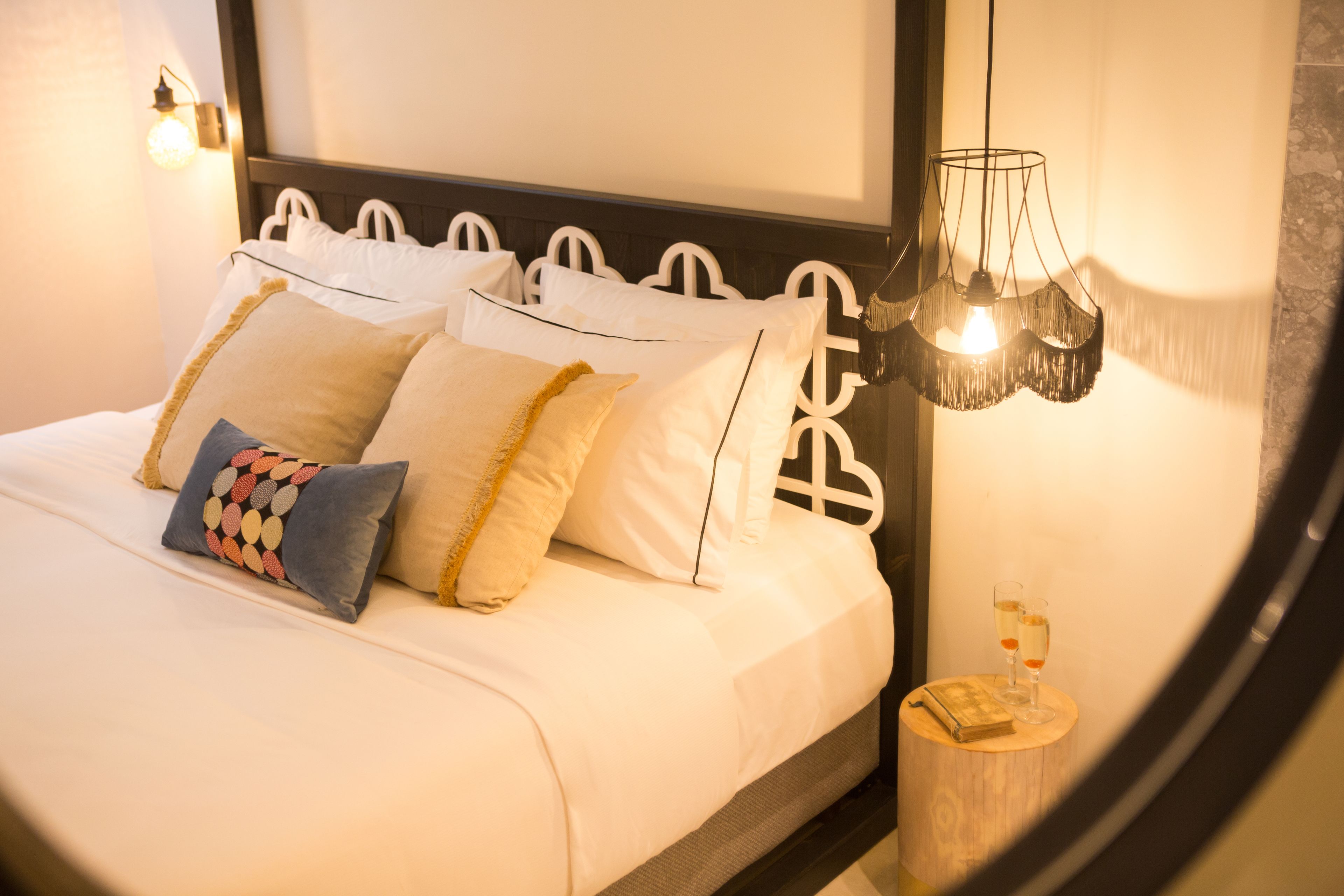 Una cama bien hecha con ropa de cama blanca y almohadones decorativos, una mesita de noche con un libro y gafas, y una lámpara de pared.