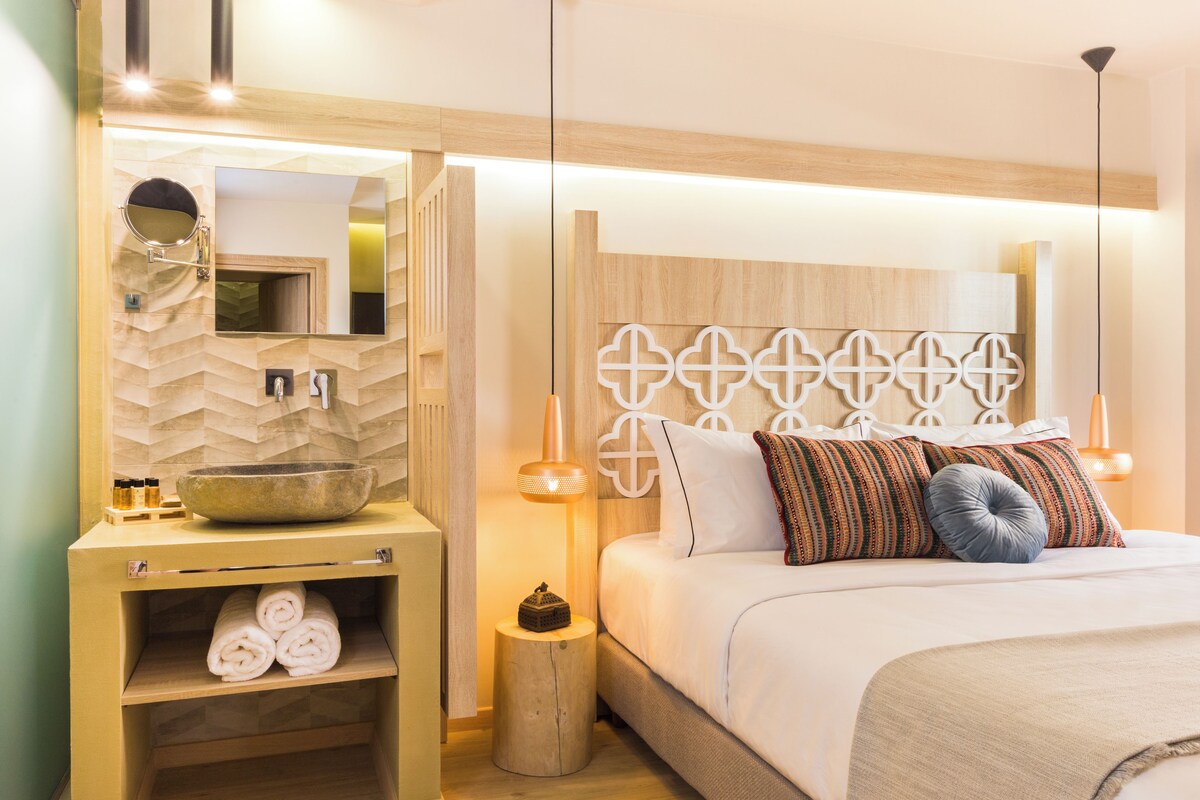 A modern hotel room with a wooden headboard, a large bed with multiple pillows, a bedside table with a bowl, and a mirror.