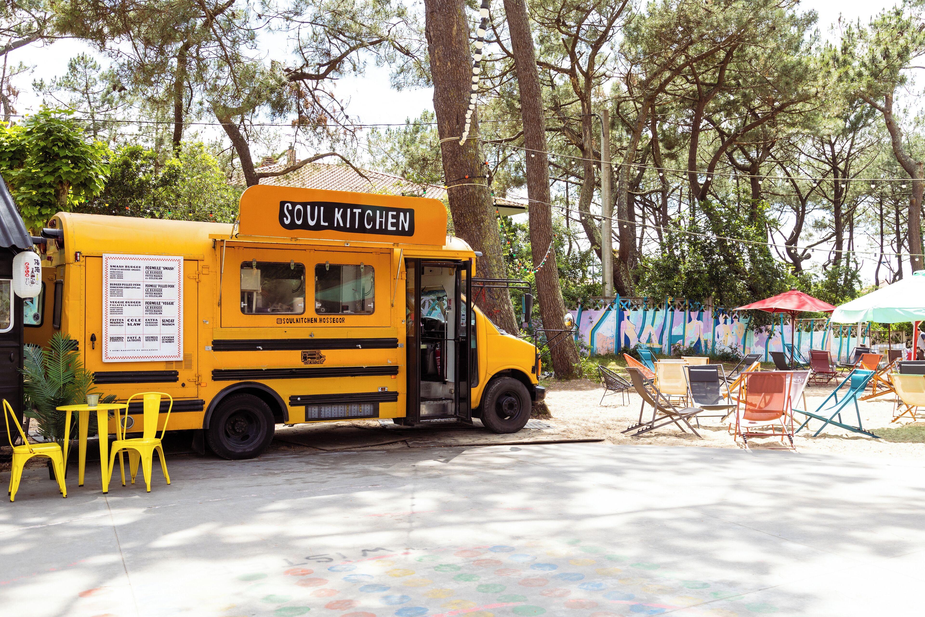 Um food truck amarelo chamado 'Soul Kitchen' estacionado em área externa arborizada, com cadeiras e mesas coloridas.