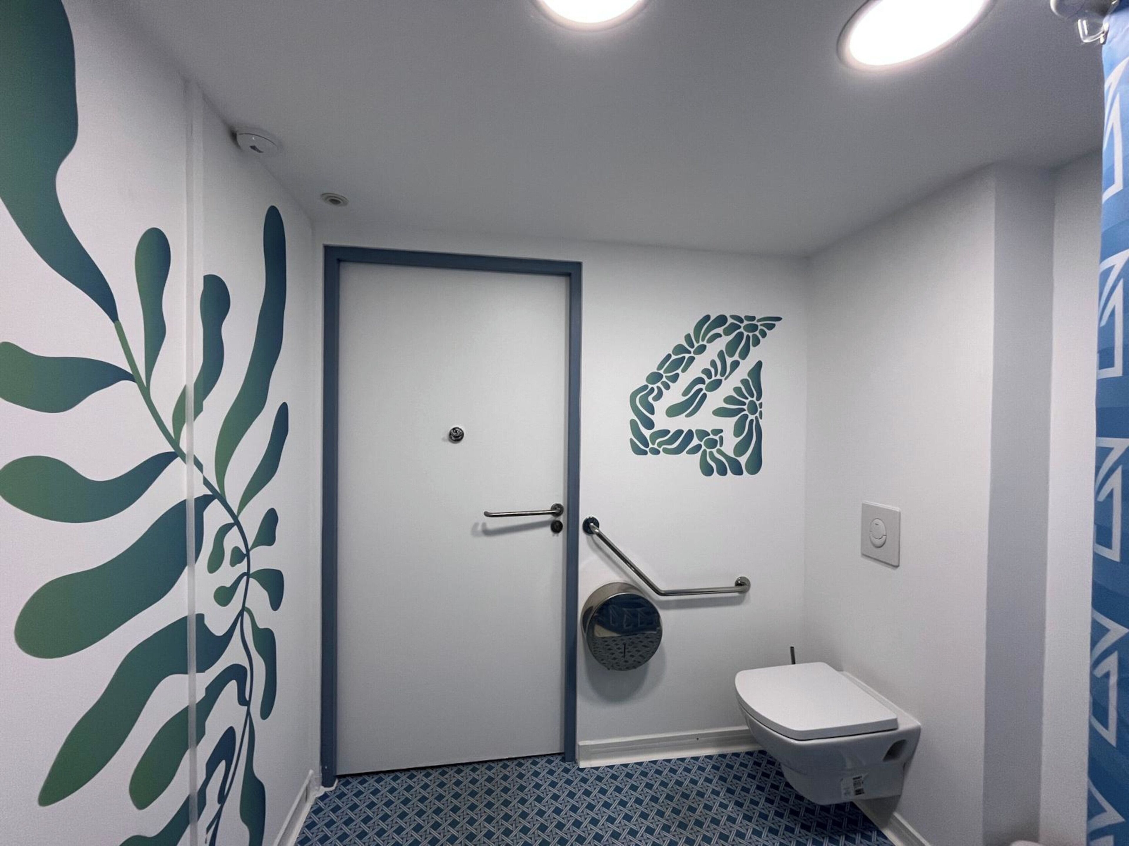 A small bathroom with a white door, a toilet, a handrail, and a wall decal.