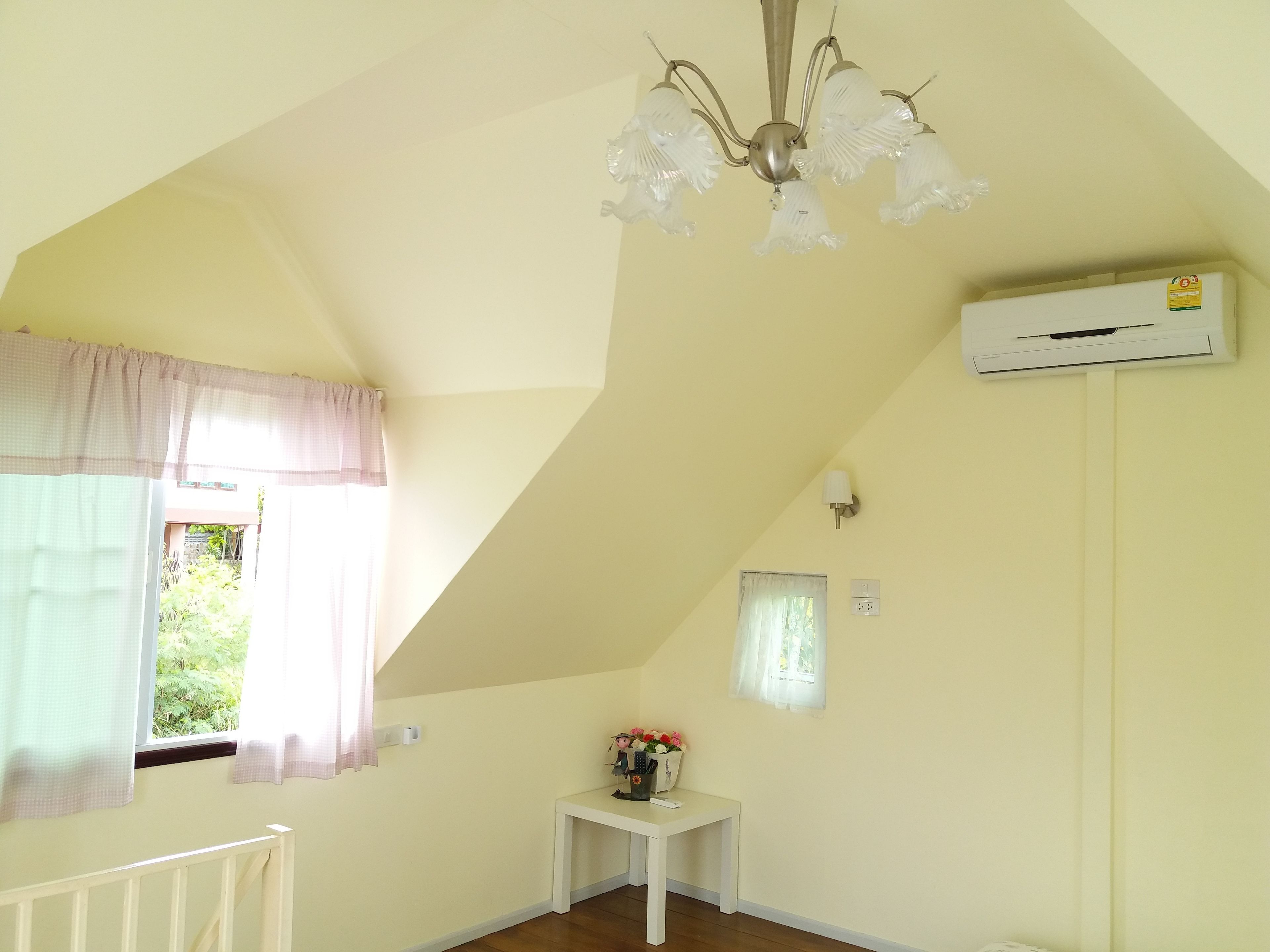 Una habitación con techo inclinado, un radiador blanco, una pequeña mesa con un florero y una ventana con cortinas.