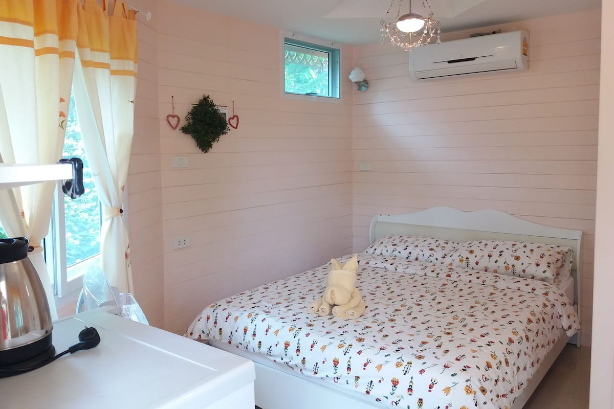 A bedroom with a bed, a window with curtains, an air conditioner, and a wall-mounted plant.