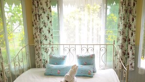 A bed with floral-patterned pillows and a Hello Kitty plush toy, a window with floral curtains, and a metal bed frame. - Hello Sunny Home (Pak Chong)