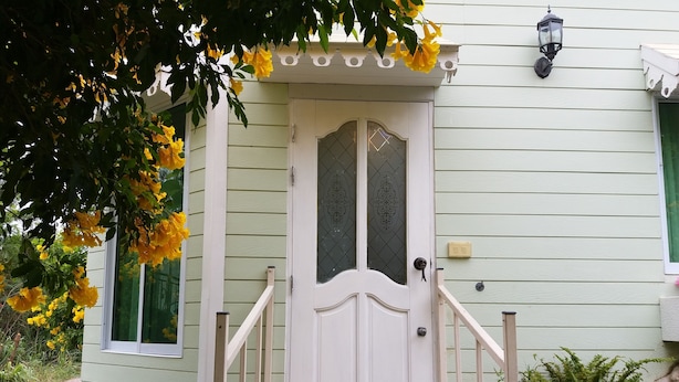 A front view of a house with a white door, a small porch with a wooden railing, and a window with a decorative glass panel. - Hello Sunny Home (Pak Chong)