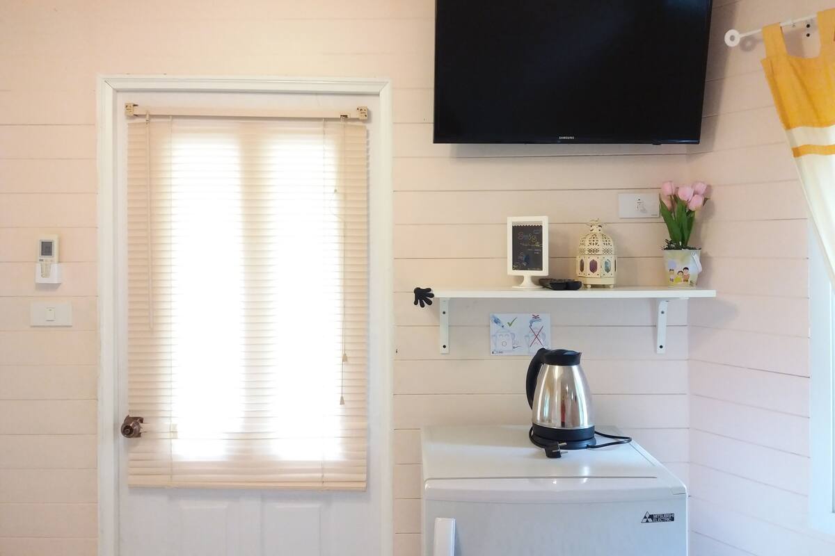 A room with a white door, a television mounted on the wall, a small shelf with a picture frame, a kettle, and a potted plant.