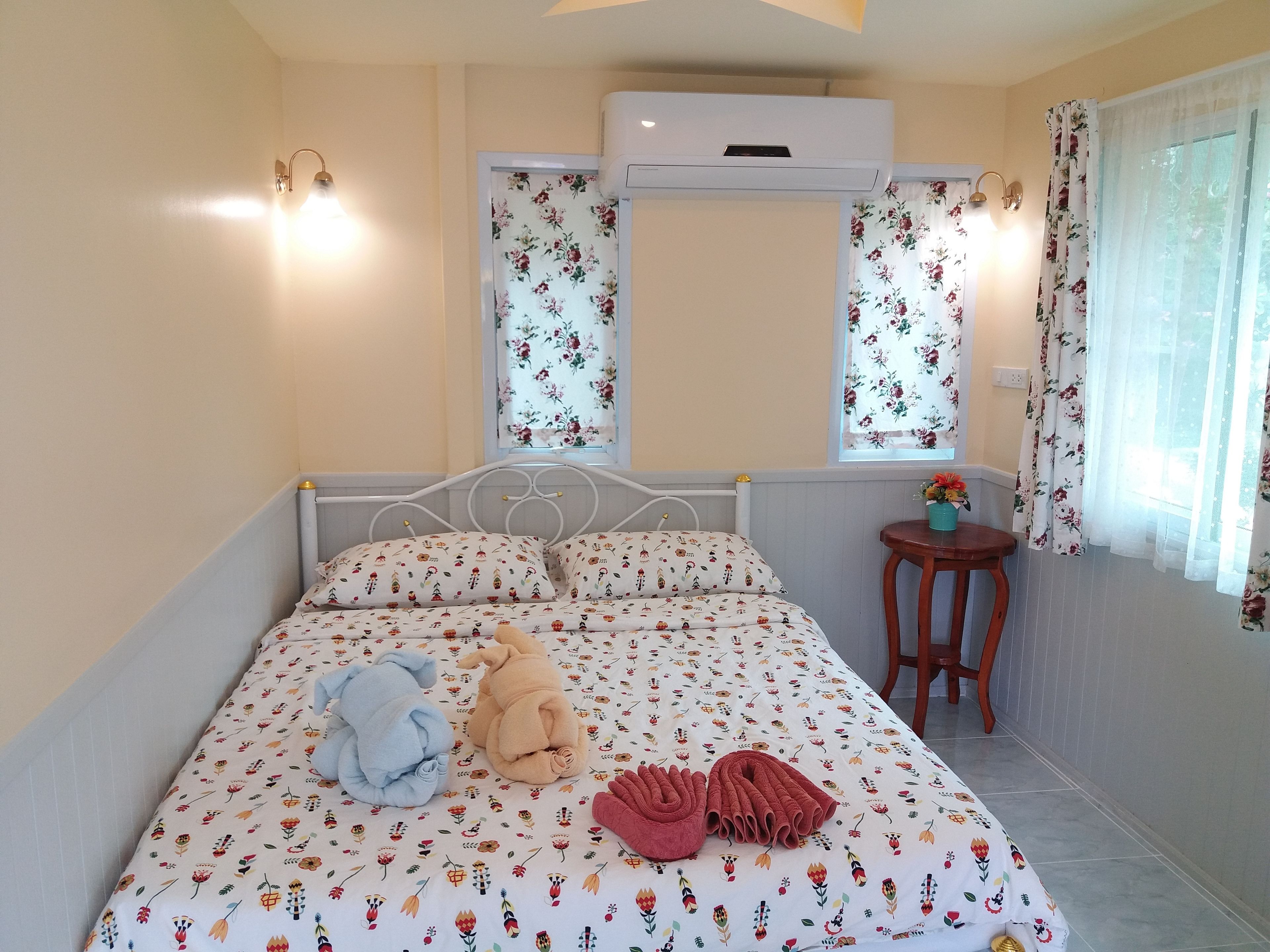Un dormitorio con cama, cortinas florales, una mesita de noche con planta y un equipo de aire acondicionado.