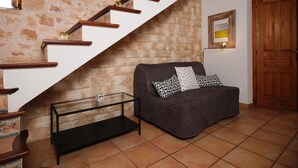 A staircase with wooden steps and a white wall with a stone pattern. There is a dark-colored sofa with patterned cushions and a small table with a glass top. A wooden door and a framed picture are visible in the background. - S'hort den Carrio (Ses Salines)