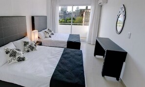 A modern hotel room with two beds, a nightstand, a bench, and a view of the city. - Lofts & Suites Rosario (Rosario)