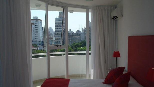 A hotel room with a bed, red pillows, a red lamp, and a view of a cityscape through a large window. - Lofts & Suites Rosario (Rosario)