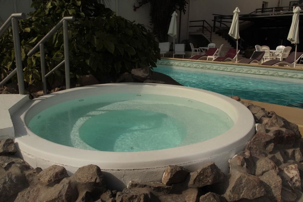 Un jacuzzi rodeado de rocas, con una piscina y sillones de descanso al fondo.