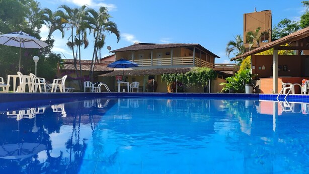 A swimming pool with lounge chairs and umbrellas, surrounded by buildings and palm trees. - Hotel Pousada Caminho da Praia (Guarapari)