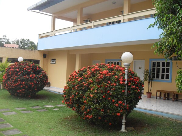 A building with a blue upper floor and a yellow lower floor, surrounded by well-maintained hedges with red flowers. - Hotel Pousada Caminho da Praia (Guarapari)