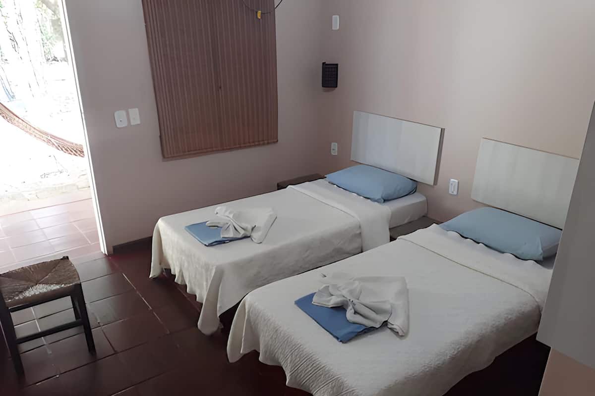 Hotellihuoneessa on kaksi erillistä sänkyä, joissa on valkoinen peitto ja sininen tyyny. Huoneen nurkassa on puinen tuoli ja pieni pöytä.