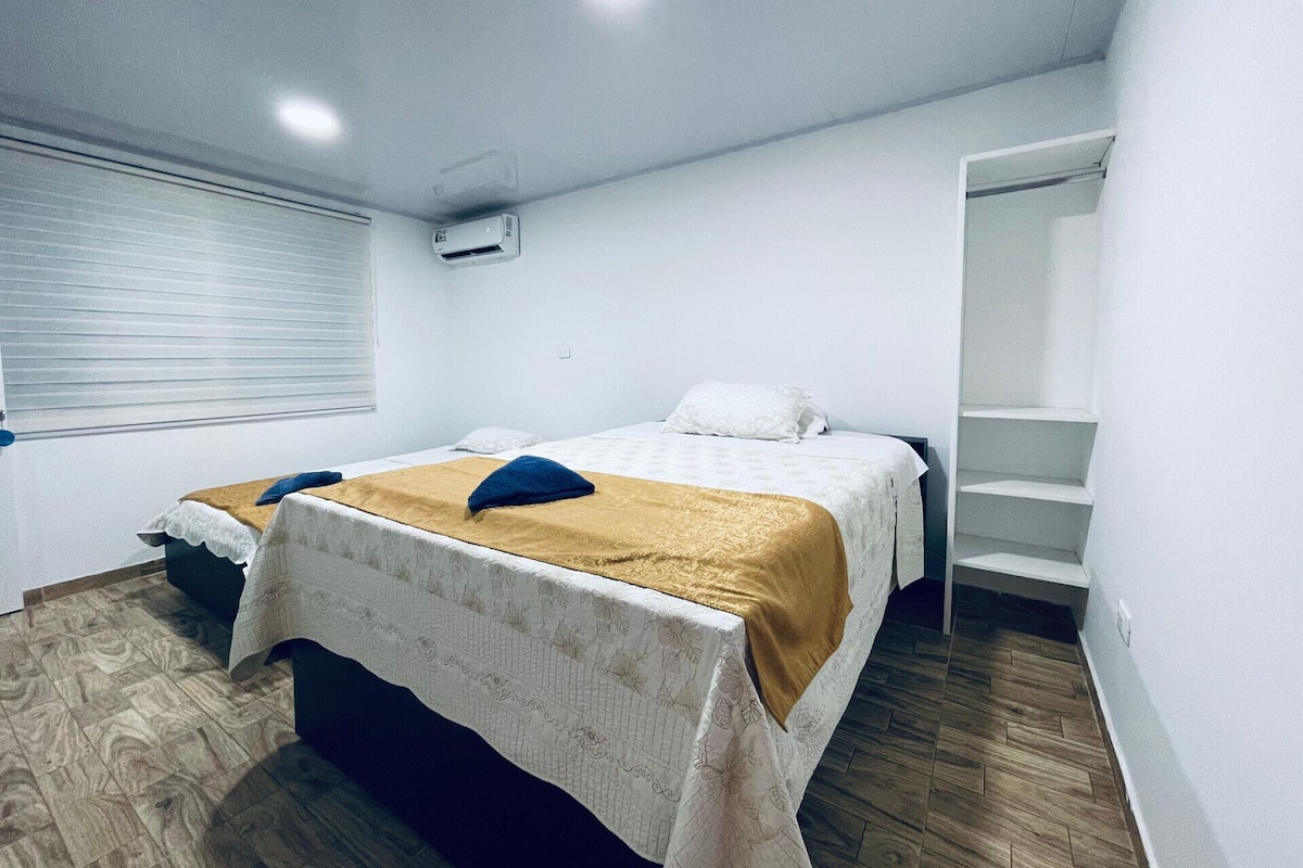 A bedroom with a bed, a window with blinds, a white air conditioning unit, and a staircase.