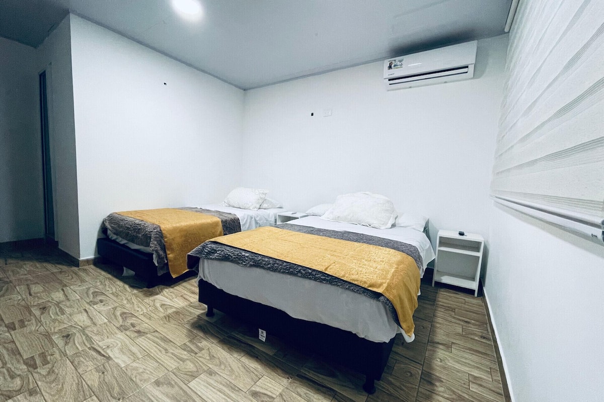 A room with two beds, a white air conditioning unit, a small bedside table, and a wooden floor.