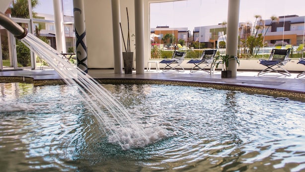 A pool area with water jets, lounge chairs, and a view of buildings through large windows. - Alto Miramar Resort & Spa (Miramar)