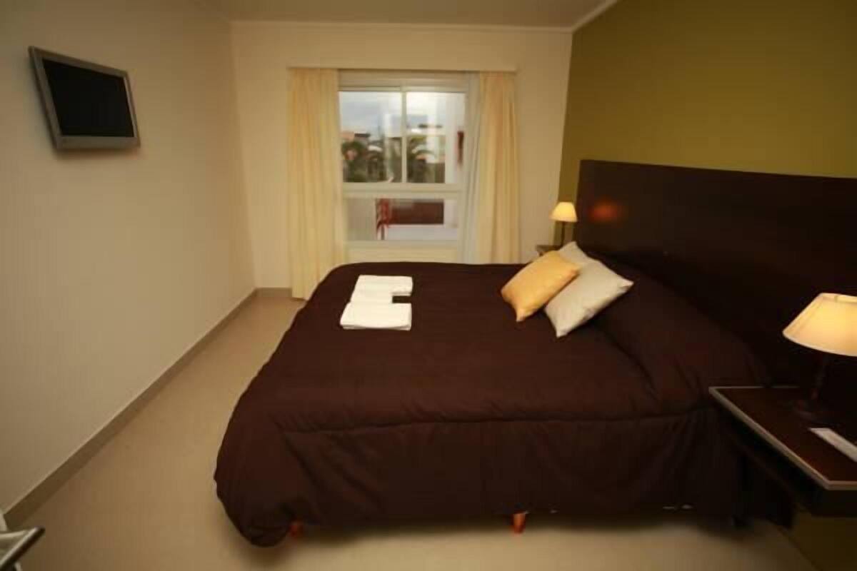 A hotel room with a large bed, a window with curtains, a bedside table with a lamp, and a television on the wall.