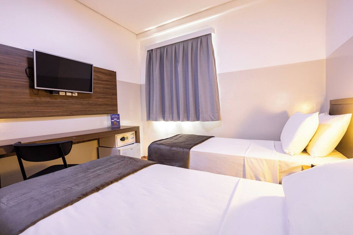 A hotel room with two beds, a desk, a television, and a window with curtains.