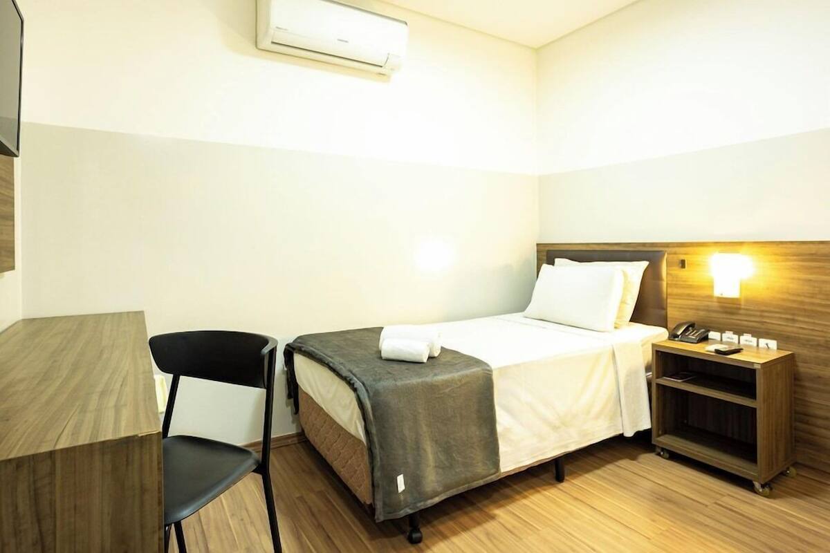 A hotel room with a bed, a desk, a chair, a nightstand, and an air conditioning unit.
