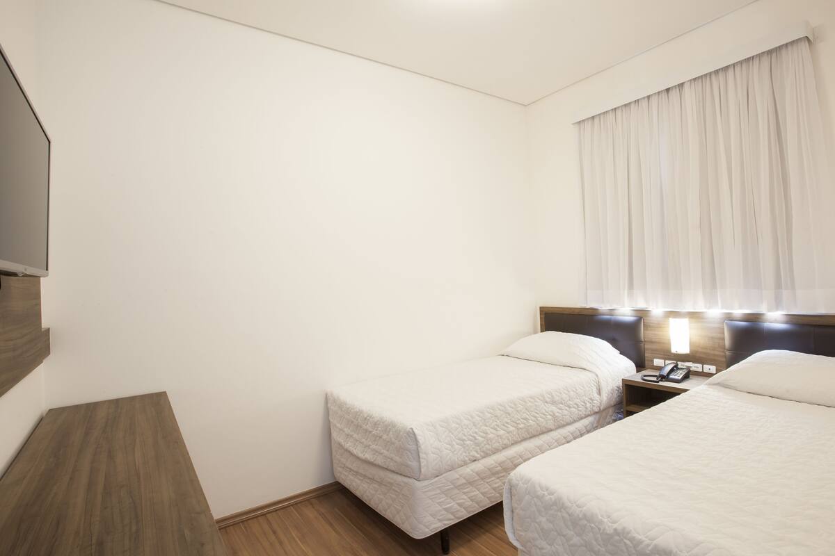 A hotel room with two beds, a bedside table with a lamp, a phone, and a TV mounted on the wall.