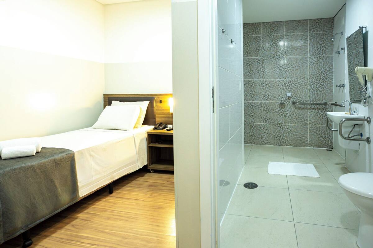 A hotel room with a bed, bedside table, and a bathroom with a shower, sink, and toilet.