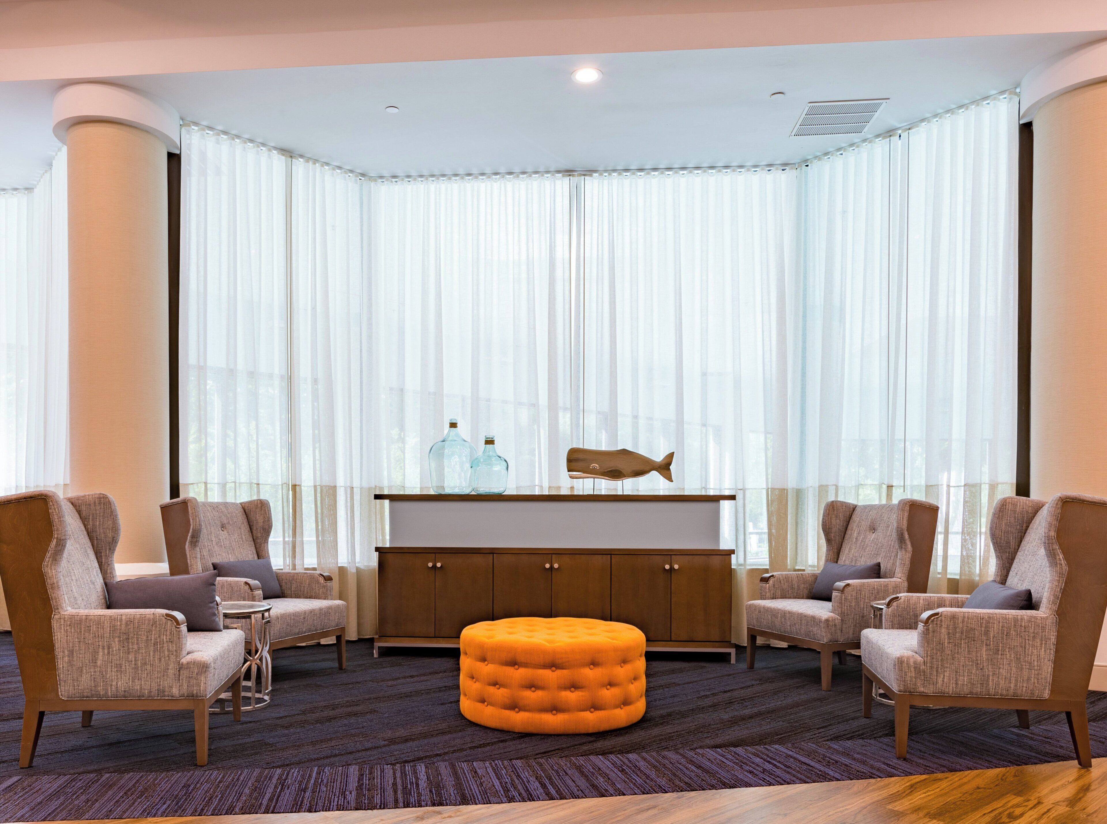 A modern living room with a dark carpet, two armchairs, a wooden ottoman, a glass cabinet with decorative items, and sheer curtains.