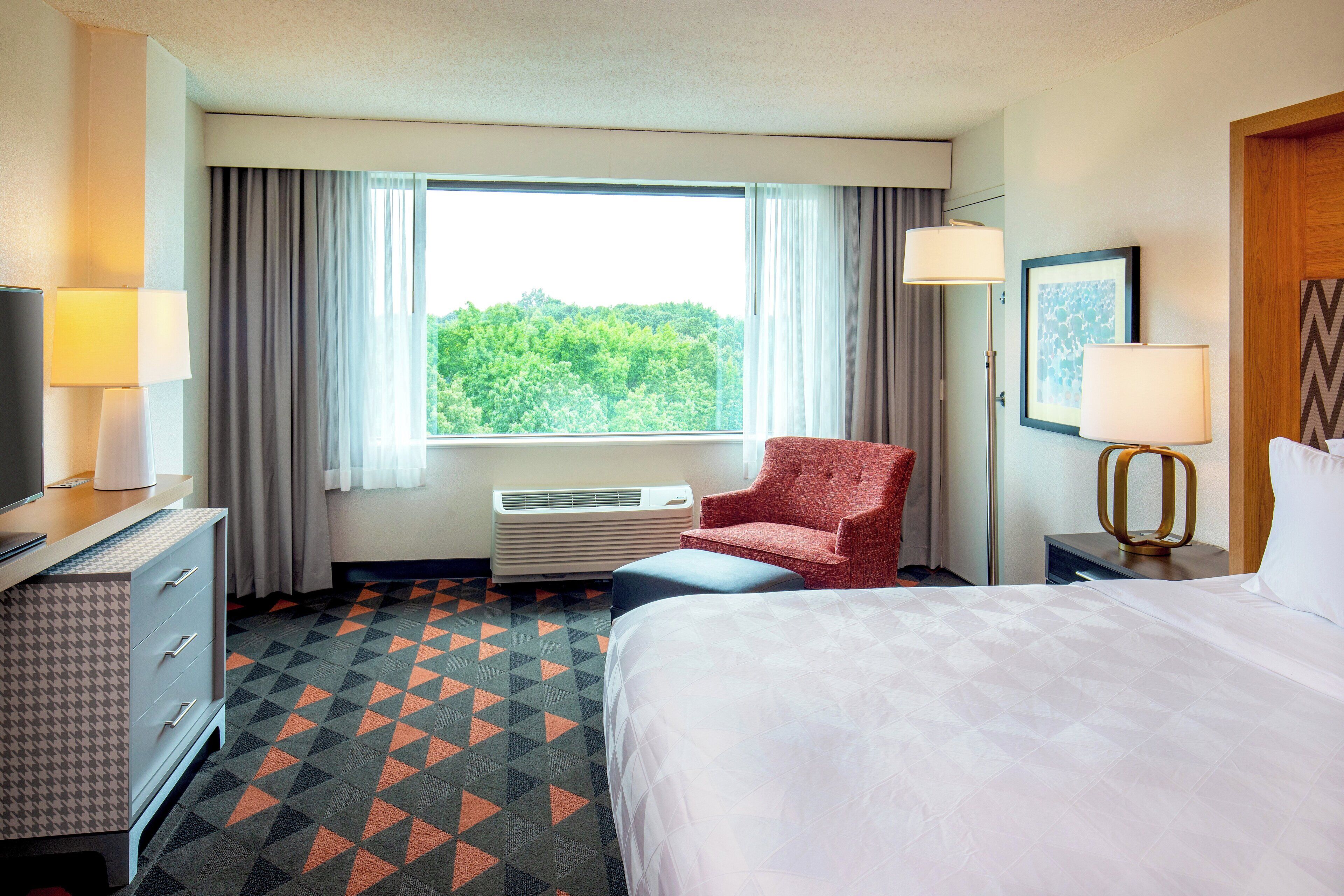A hotel room with a bed, a chair, a desk with a lamp, a TV, and a window with curtains.