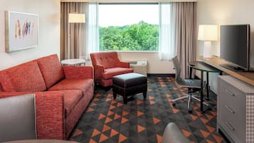 Et hotelværelse med sofa, lænestol, skammel, skrivebord, fjernsyn og et vindue med udsigt til træer.