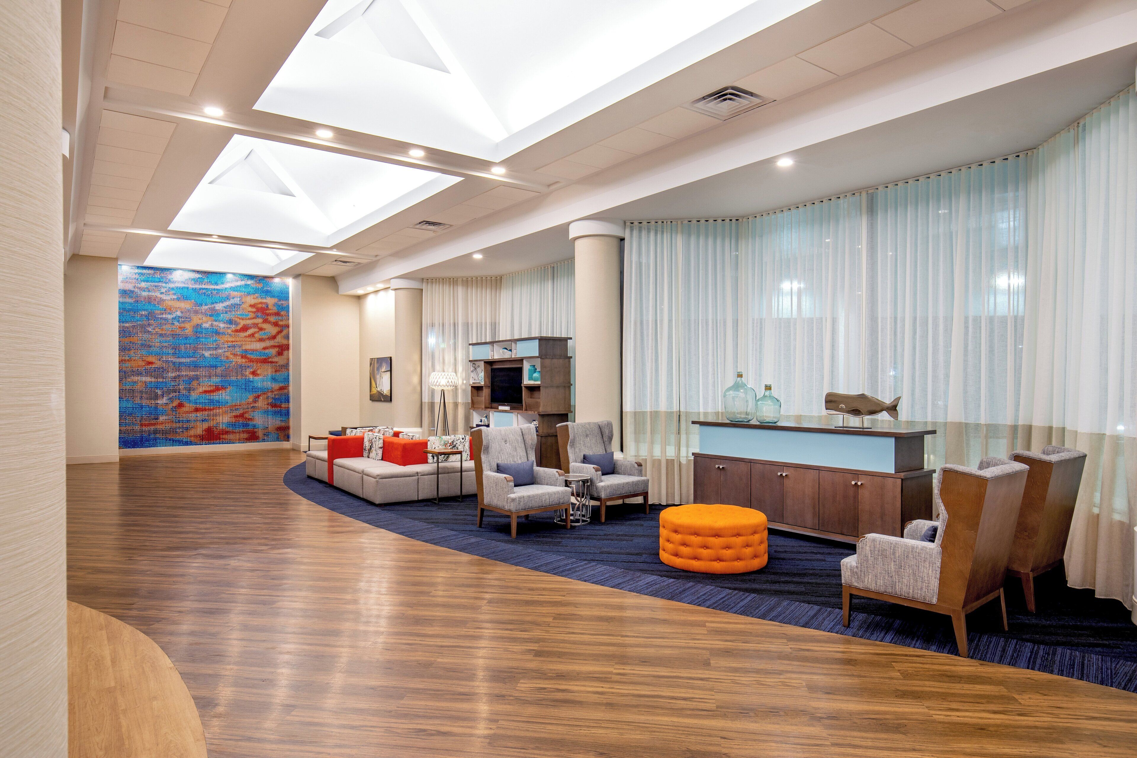 Un amplio vestíbulo de hotel con techo alto, un gran mural abstracto y colorido, un área de descanso con sofás y sillones, un mostrador de recepción de madera y un ventanal con cortinas traslúcidas.