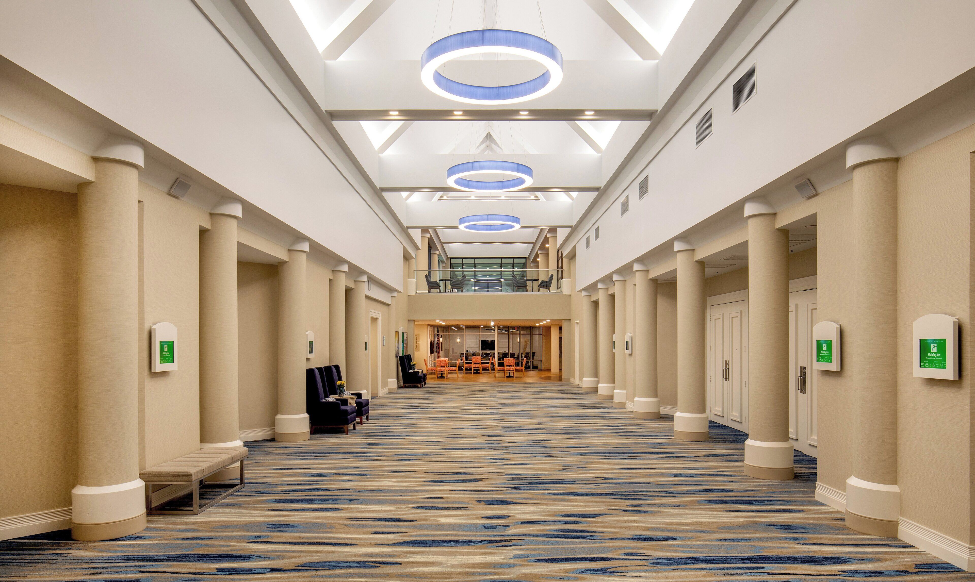 A hotel corridor with a patterned carpet, columns, and a seating area with chairs.