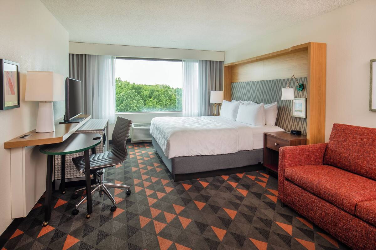 Habitación de hotel con una cama, un escritorio con computadora, televisor, sofá rojo y ventana con cortinas.