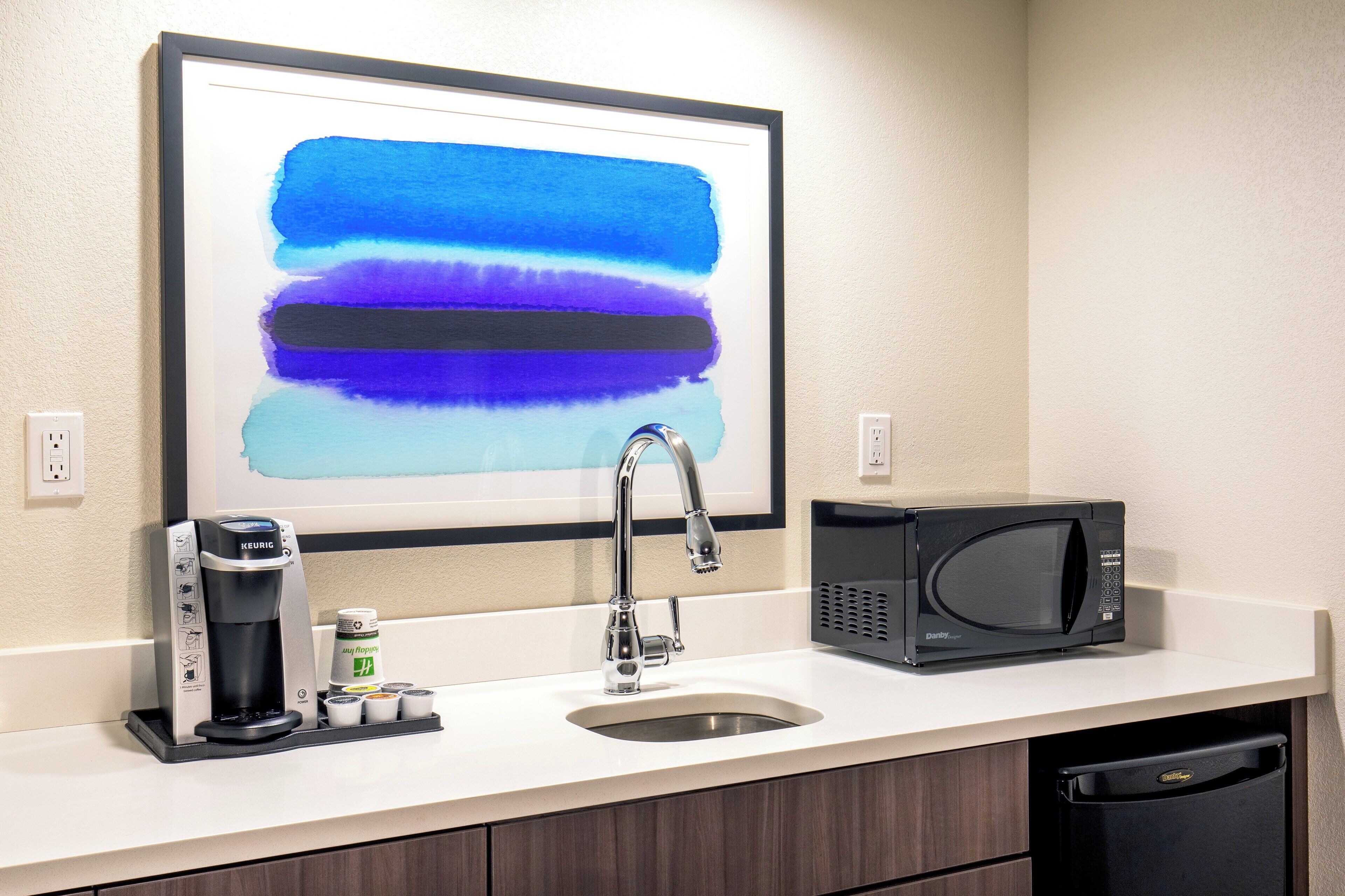 A kitchenette with a coffee maker, microwave, sink, and a framed abstract painting above the sink.