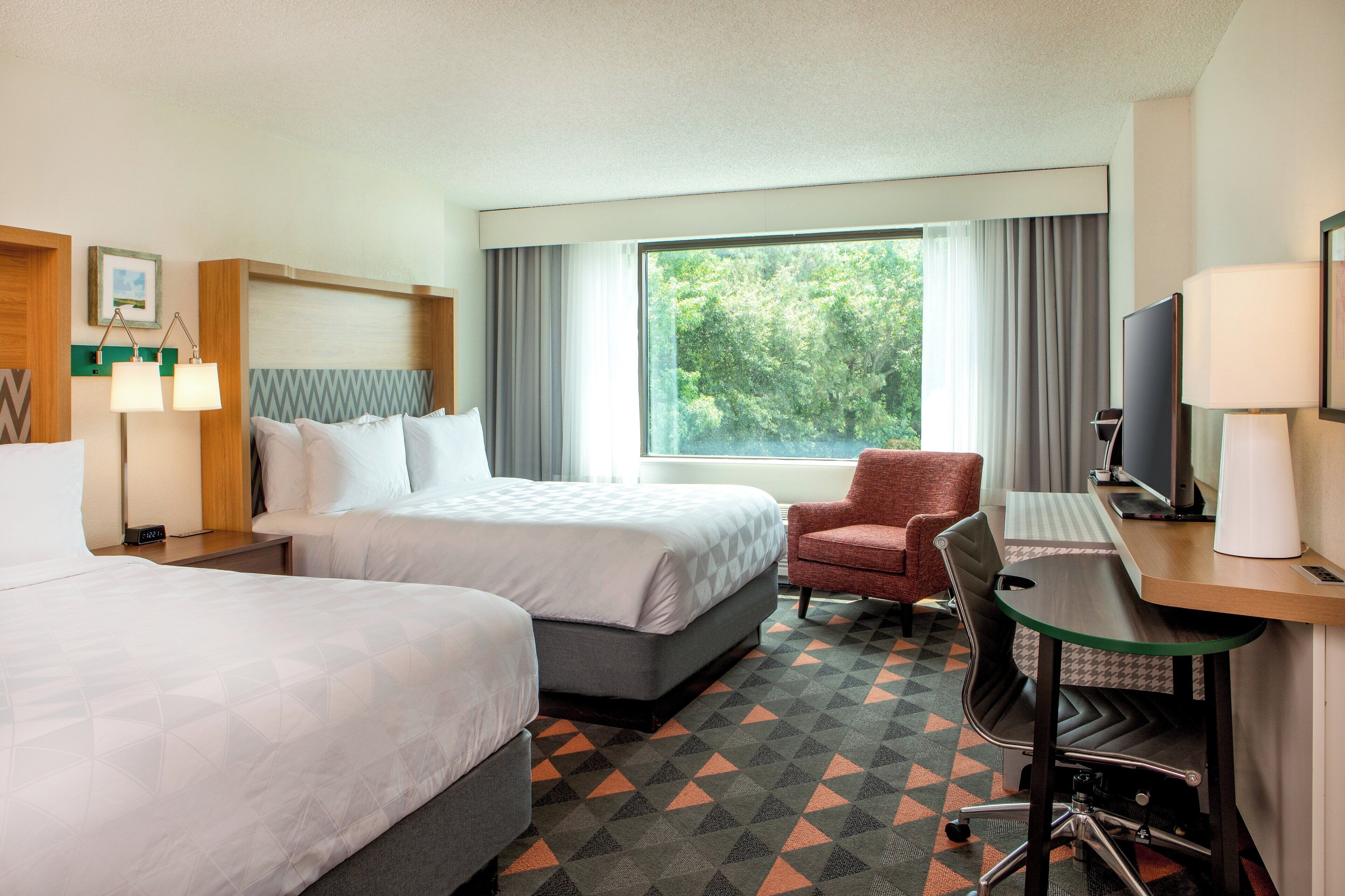 A hotel room with two beds, a desk with a chair, a TV, and a window with curtains.