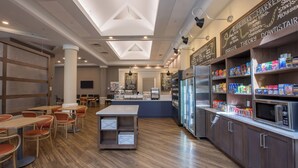 A modern café interior with wooden flooring, tables, chairs, a central island, a refrigerator, and shelves stocked with snacks. - Holiday Inn Newport News - Hampton by IHG (Newport News)