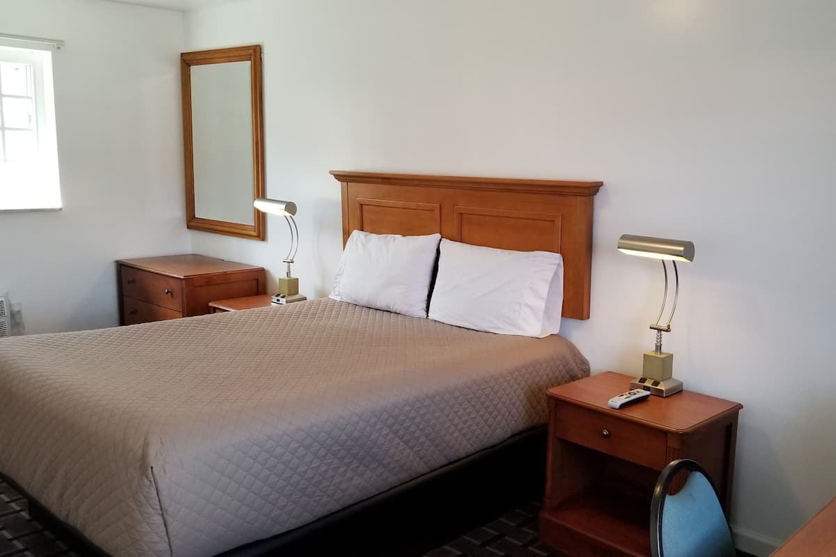 A hotel room with a large bed, two bedside tables with lamps, a wooden headboard, a mirror, a small desk with a chair, and a television.