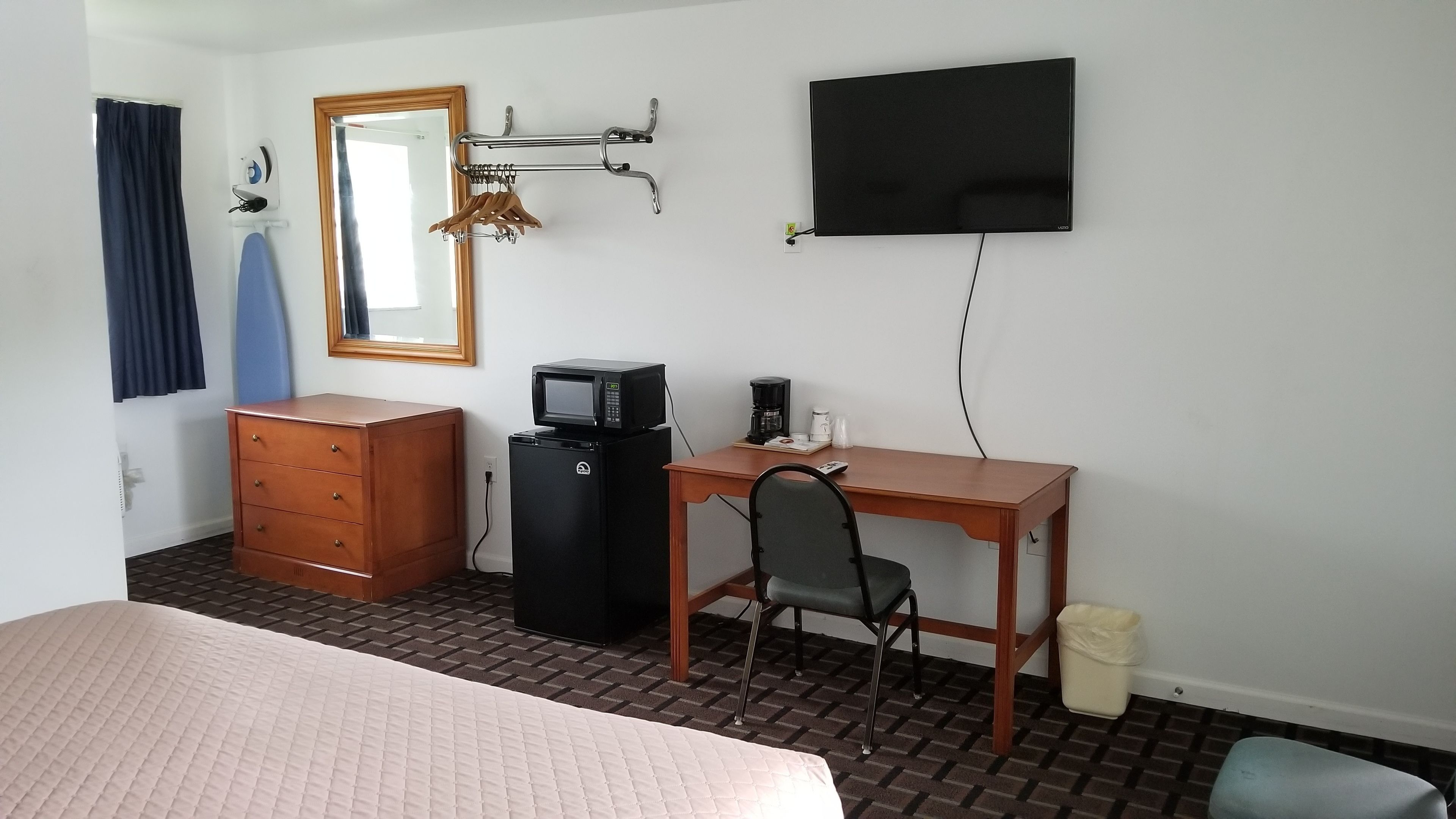 A hotel room with a bed, a wooden dresser, a desk with a chair, a television, a microwave, a coffee maker, and a trash bin.