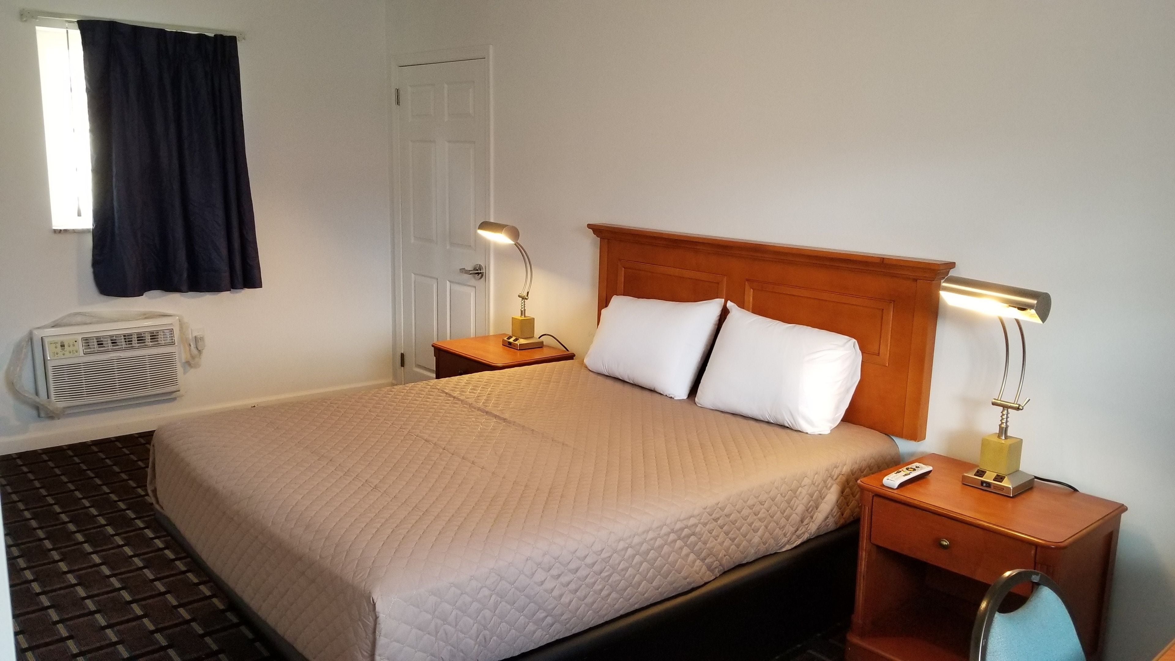 A hotel room with a bed, two bedside tables with lamps, a heater, a window with curtains, and a door.