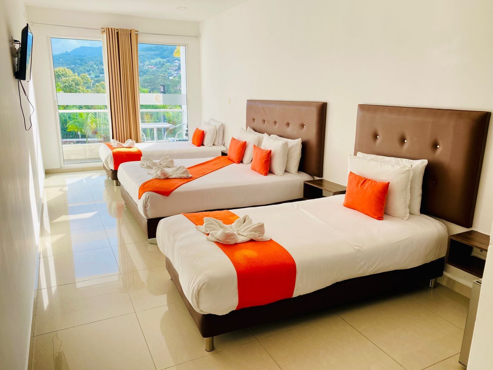 A hotel room with three beds, a television, and a balcony with a view of greenery.