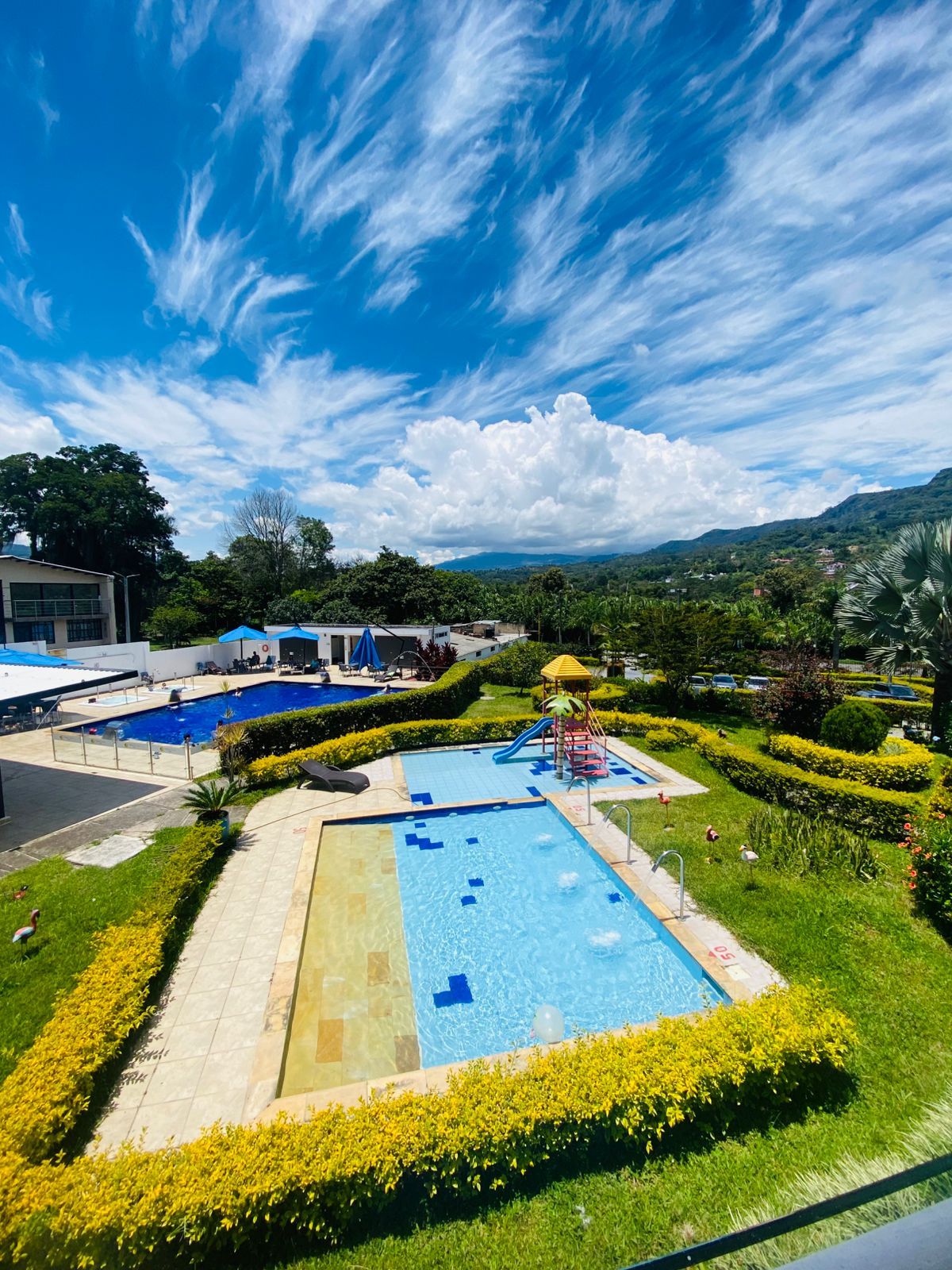 A swimming pool area with multiple pools, surrounded by well-maintained gardens and a view of distant hills.