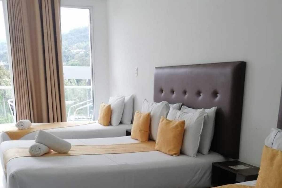 Une chambre d’hôtel avec deux lits, chacun agrémenté de draps blancs et d’un plaid marron. Une grande fenêtre, ornée de rideaux, donne sur la pièce, qui présente un design moderne avec une tête de lit sombre.