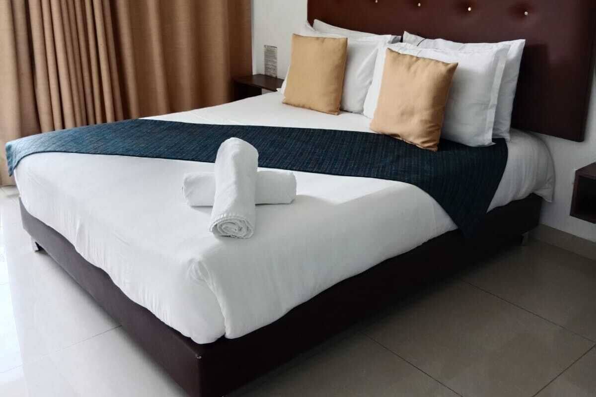 Hotellihuone, jossa on suuri sänky, tumma pääty, valkoiset vuodevaatteet ja sängyllä rullalle taitettu pyyhe.