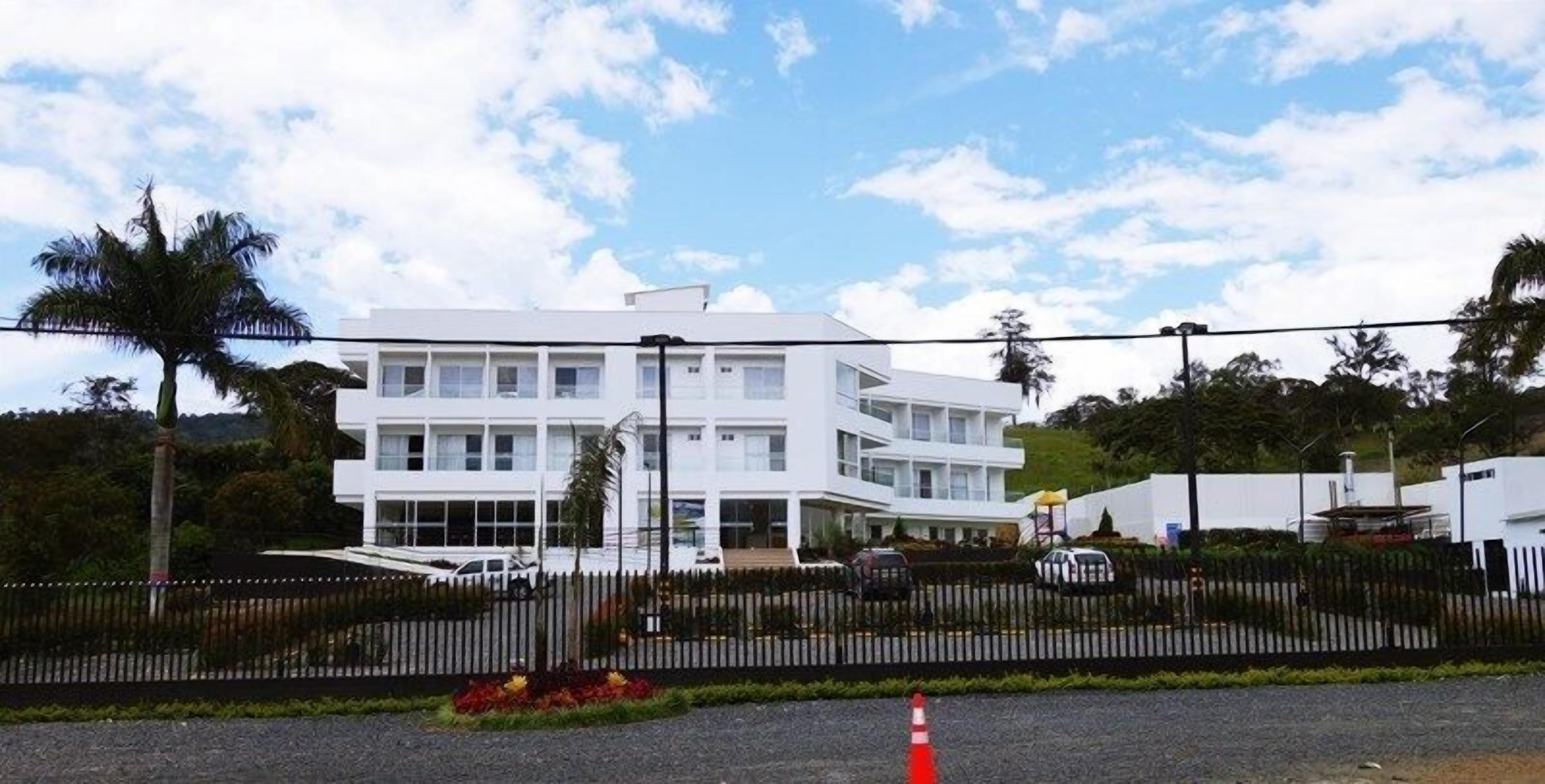 A modern white building with multiple windows and a flat roof, surrounded by a black fence and palm trees.