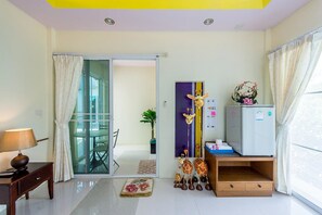 Quarto compacto com porta de vidro deslizante para uma varanda, uma pequena mesa com abajur, um refrigerador e uma escultura decorativa de girafa.