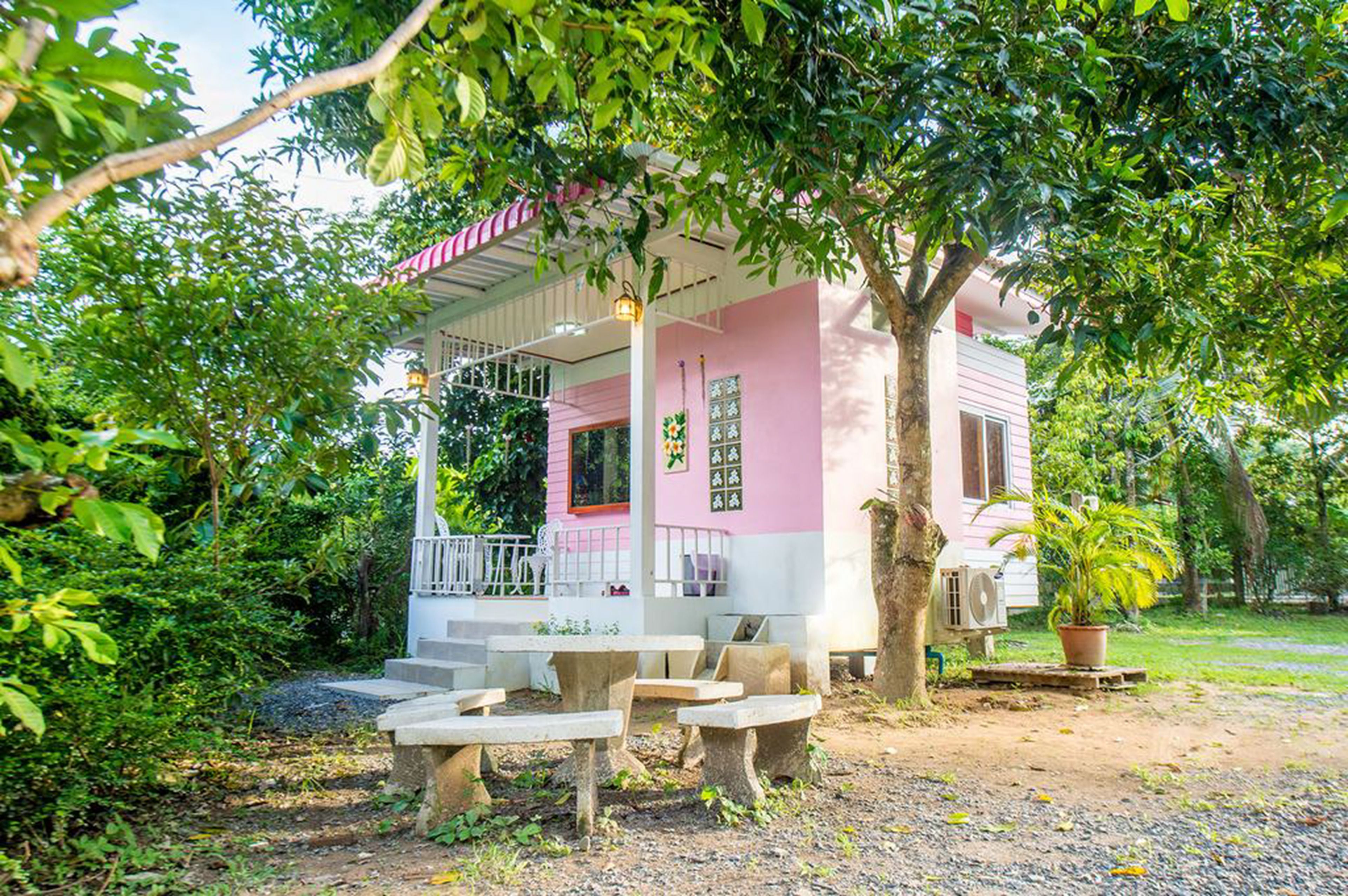 Un bâtiment rose et blanc avec un porche abrité, des sièges à l’extérieur et un arbre au premier plan.