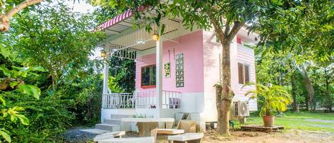 Un bâtiment rose et blanc avec un porche abrité, des sièges à l’extérieur et un arbre au premier plan.