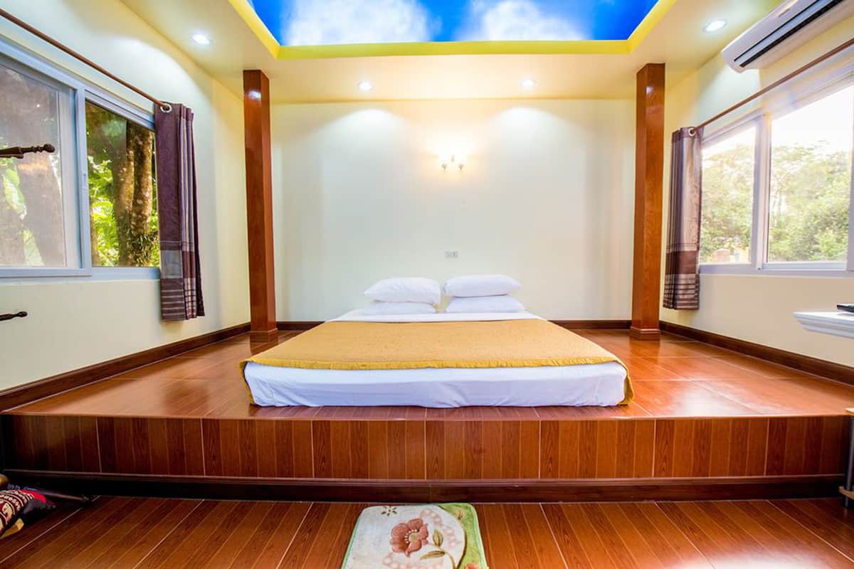 A bedroom with a bed centered on a wooden platform, large windows on both sides, and a ceiling with a blue sky design.