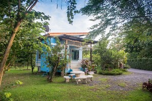 Uma pequena casa azul com varanda e área de estar externa, cercada por vegetação exuberante.